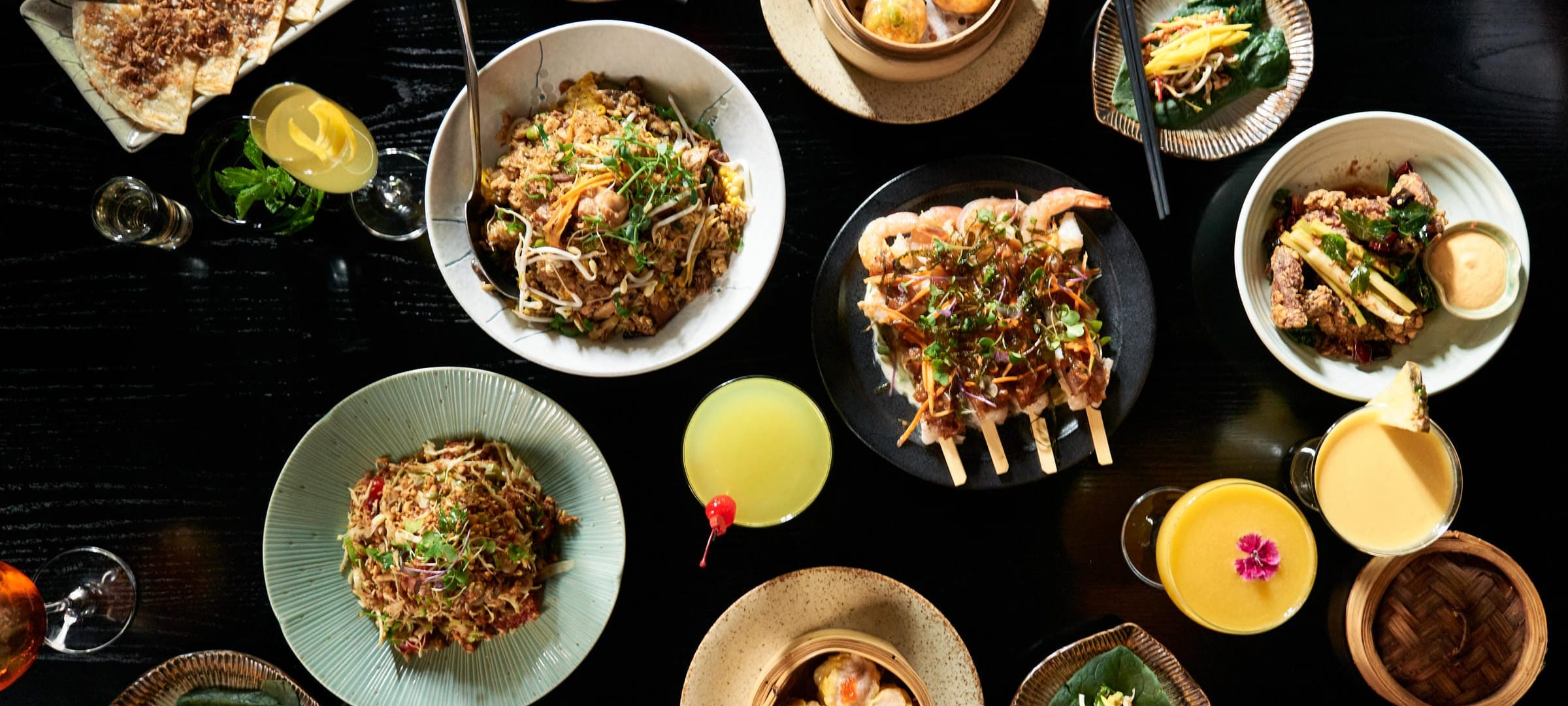 A table of Zodiac dishes to share, seen from above: noodles, seafood skewers, dumplings and cocktails