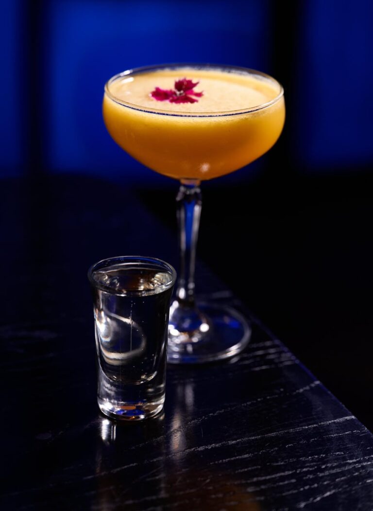 A passionfruit coupe cocktail with a flower garnish and a side shot on the blue-lit bar at Zodiac